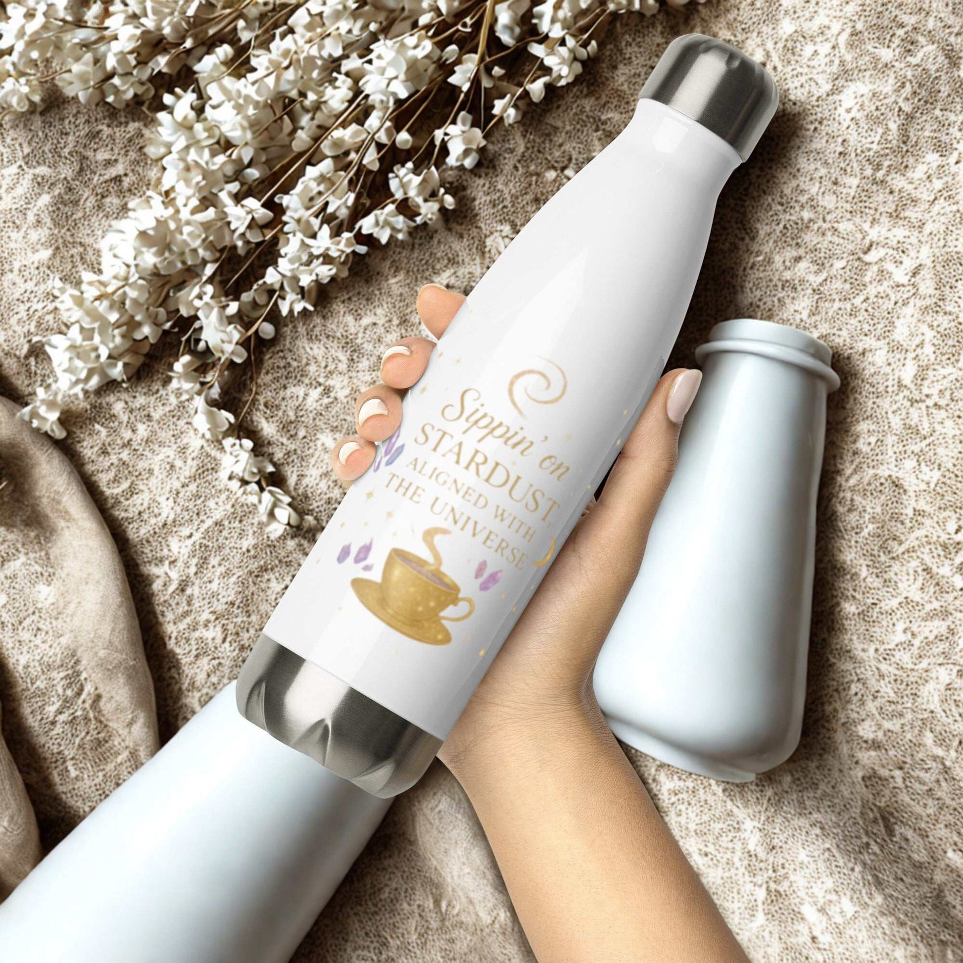 White Sippin on Stardust Water Bottle with gold text and tea cup design held in hand on beige textured background with white flowers.