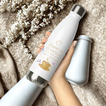 White Sippin on Stardust Water Bottle with gold text and tea cup design held in hand on beige textured background with white flowers.