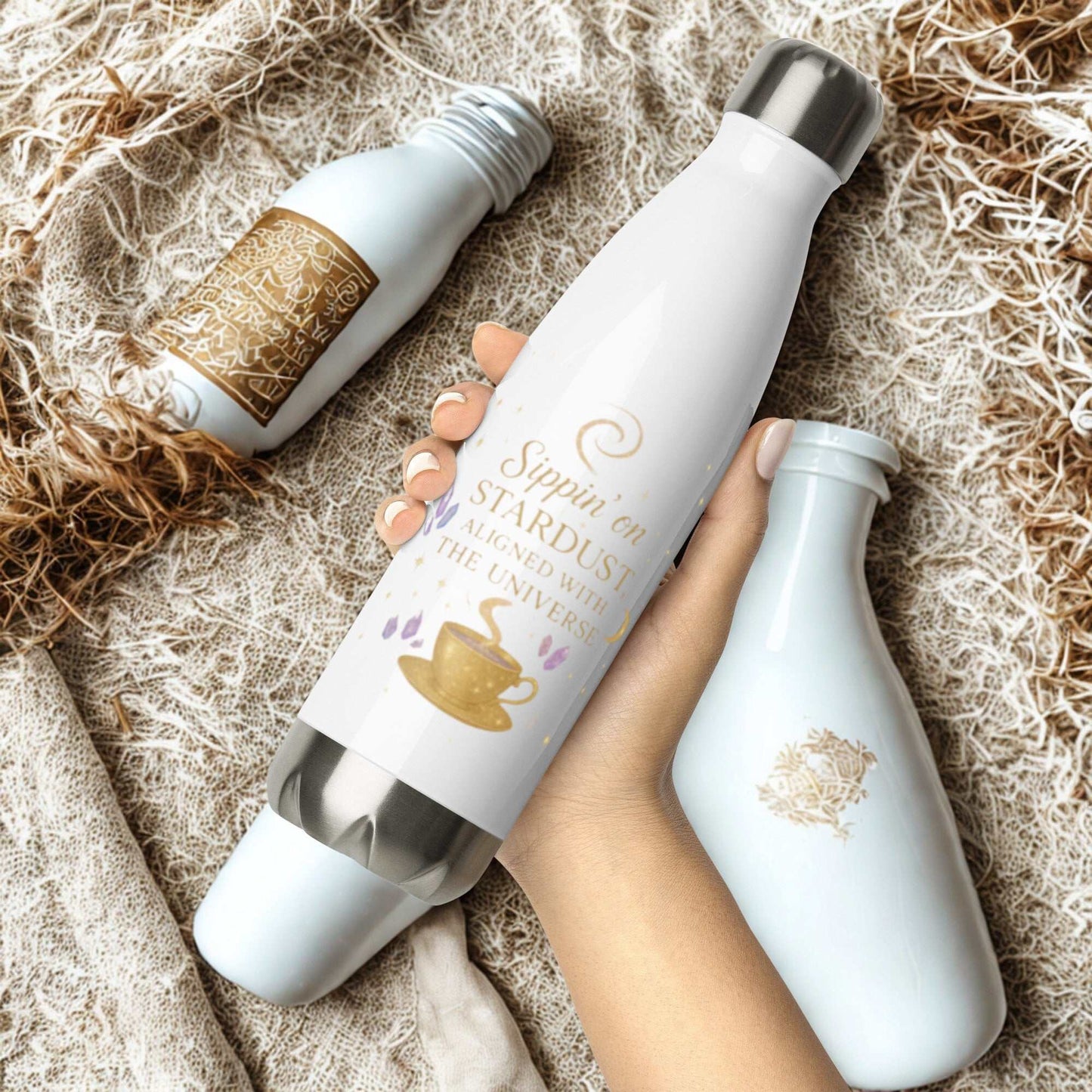 White Sippin on Stardust Water Bottle with gold design held in hand among other bottles on textured fabric background