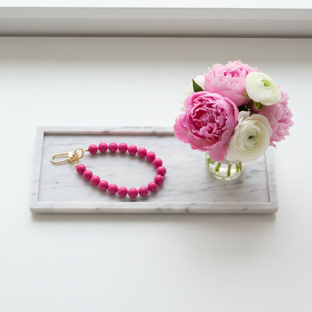Roze telefoonkoord met ronde kralen en goudkleurige clip, stijlvol gepresenteerd op een marmeren dienblad naast een boeket roze en witte bloemen.