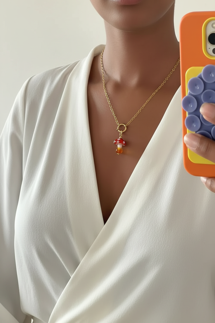 Speelse fairycore paddenstoel bedel van hars gedragen door een vrouw in witte blouse met een gekleurde telefoon in haar handen.
