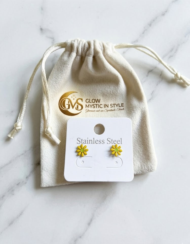 "Productfoto van Glow Mystic in Style gele bloem-oorknopjes op een wit 'Stainless Steel' kaartje. Het kaartje rust tegen een luxe, crème-wit fluwelen zakje waarop het goudkleurige logo van Glow Mystic in Style prijkt. Het geheel ligt op een lichte marmeren ondergrond met zachte grijze aders, wat een elegante en professionele uitstraling geeft aan de verpakking en het sieraad."