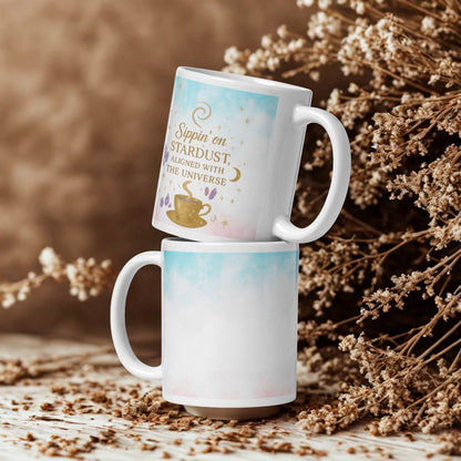 Two stacked white ceramic Sippin on Stardust Coffee Mugs with celestial design and text, placed on natural textured surface with dried flowers background