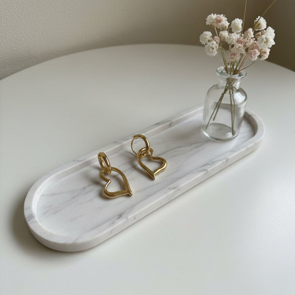Goudkleurige hartjes oorbellen op een wit marmeren dienblad naast een klein glazen vaasje met witte droogbloemen.