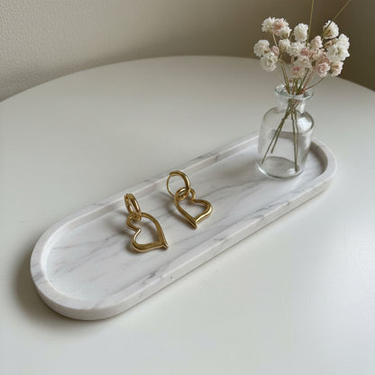 Goudkleurige hartjes oorbellen op een wit marmeren dienblad naast een klein glazen vaasje met witte droogbloemen.