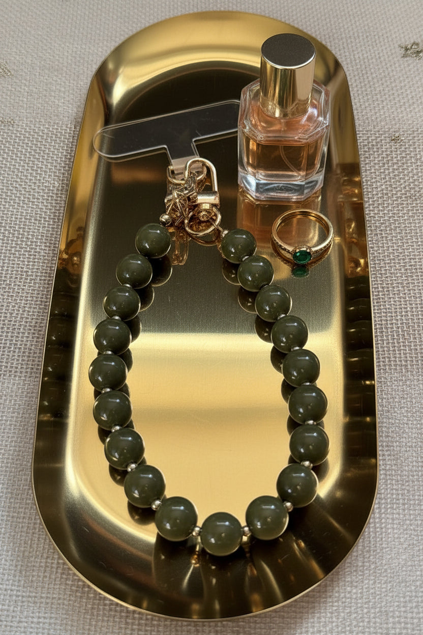 vooraanzicht van een groene kralen telefoonkoord met gouden details op een goudkleurige glanzende schaal met 2 lifestyle producten ernaast.