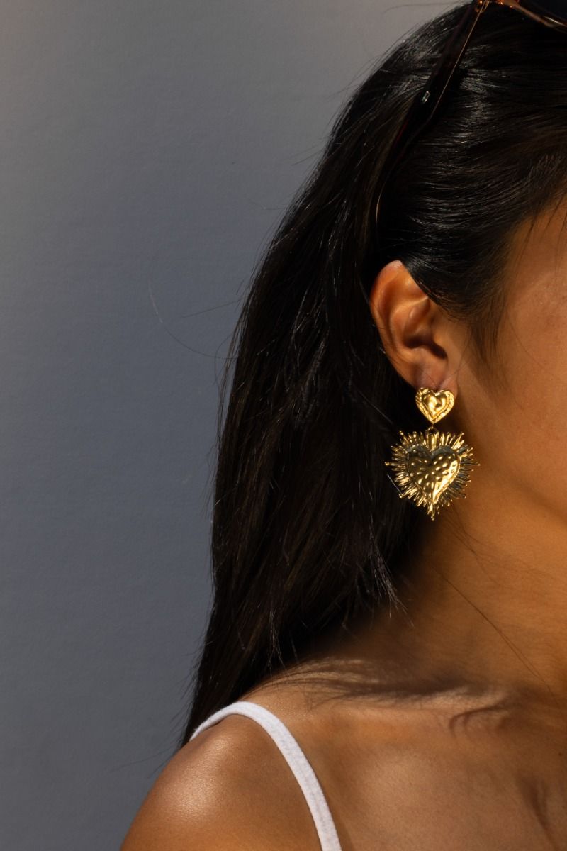 Modelfoto van grote, gouden hart oorbellen met een spiky design. Een trendy en statement sieraad gedragen door een vrouw met donker haar en een witte top.