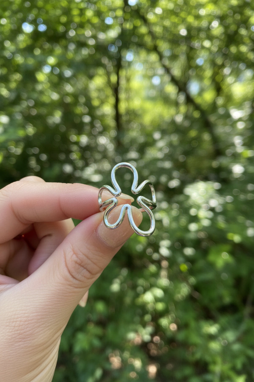 "Zilverkleurige RVS Abstract Flower ring van Glow Mystic in Style, vastgehouden tegen een natuurlijke achtergrond van groene bladeren in het daglicht."