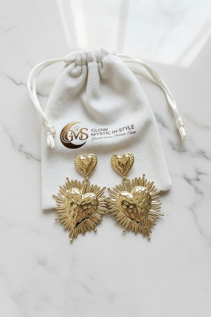 Set van twee goudkleurige RVS statement hart oorbellen gepresenteerd op een wit luxe velvet sieraadzakje met het logo van Glow Mystic in Style.