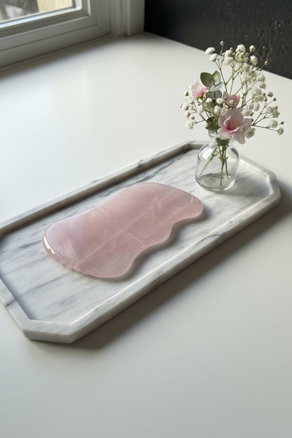 Luxe zachtroze Rozenkwarts Gua Sha massagesteen op een marmeren plateau naast een vaas met witte en roze bloemetjes- Glow Mystic in Style.