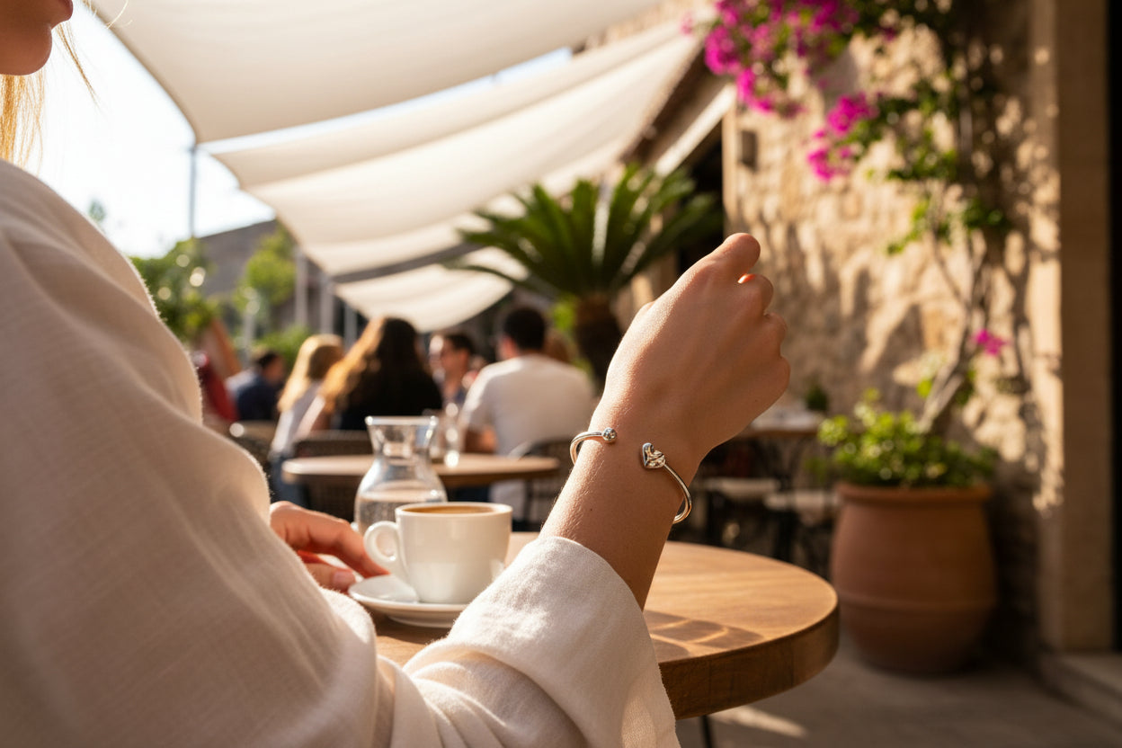 Hartbangle armband gedragen door een vrouw die op een zonnig terras zit en een kopje koffie drinkt, de achtergrond zijn meerdere mensen en bloemen en planten op het teras.