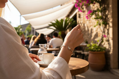 Hartbangle armband gedragen door een vrouw die op een zonnig terras zit en een kopje koffie drinkt, de achtergrond zijn meerdere mensen en bloemen en planten op het teras.