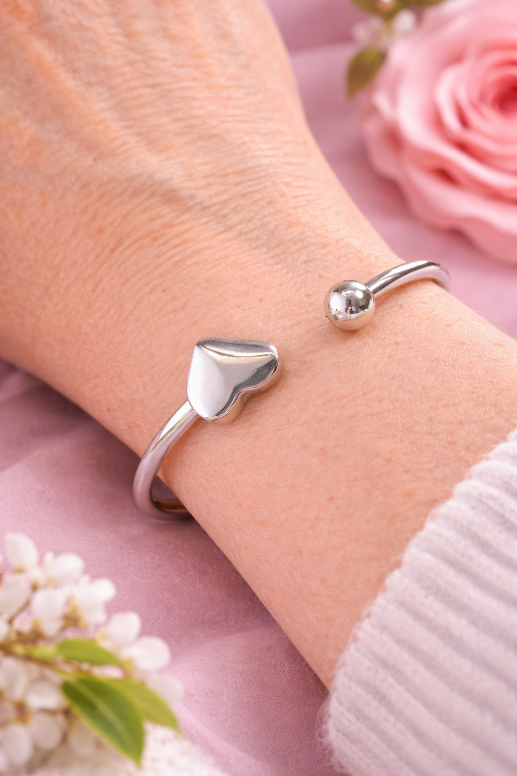 Een minimalistische zilveren hartjes-armband van Stainless Steel gedragen om een pols, gepresenteerd in een luxe setting met een roze roos en witte bloesem op een zachtroze achtergrond. One-size en licht buigbaar.
