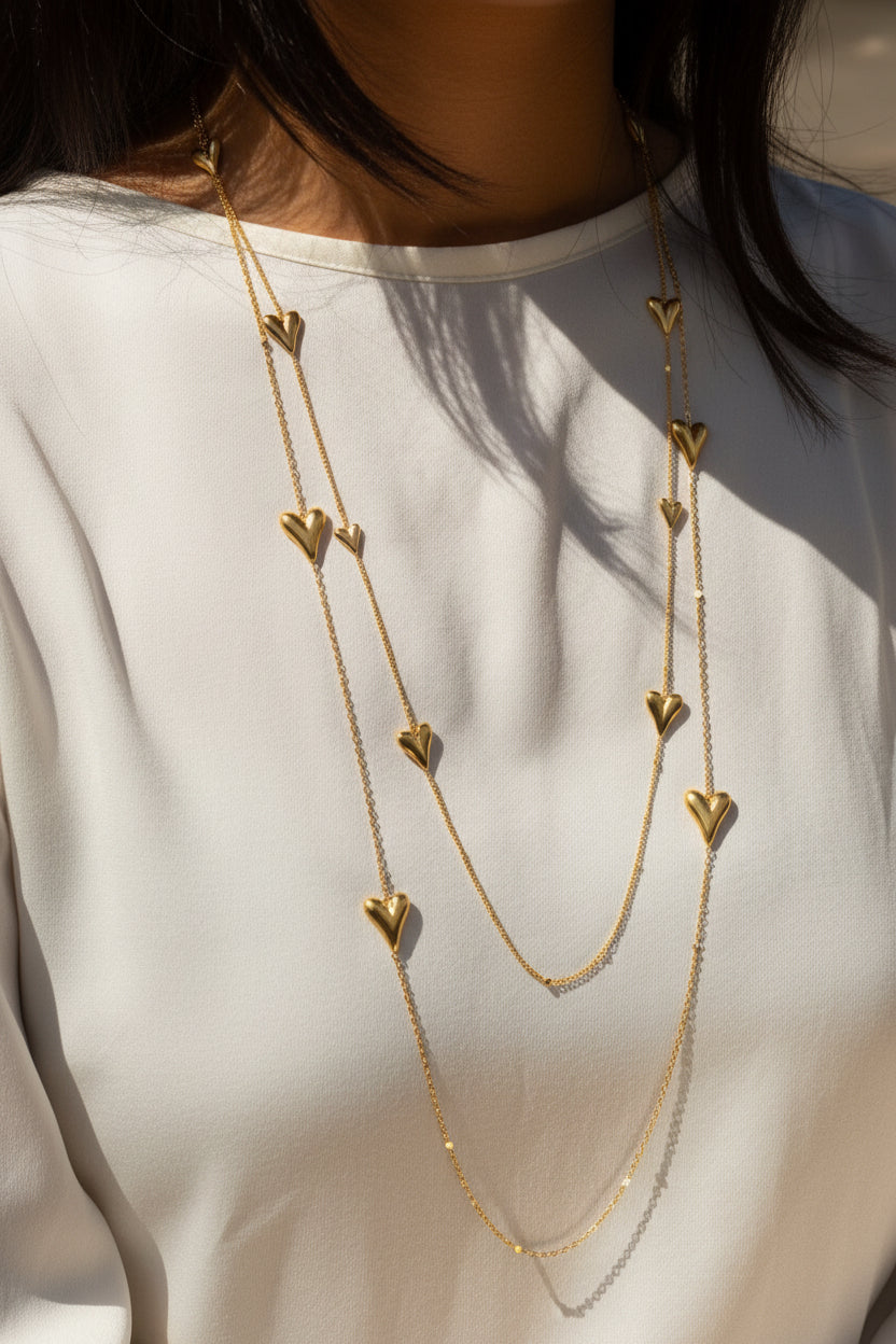 Lange goudkleurige RVS hartjes ketting dubbelgeslagen gedragen als een gelaagde set op een elegante witte blouse van een vrouw.