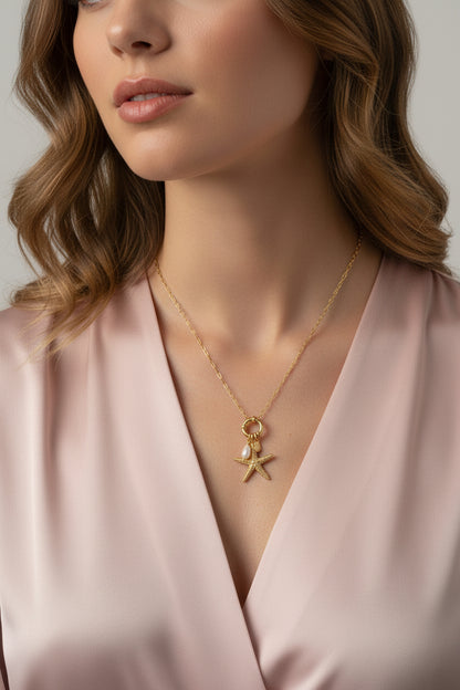 Goudkleurige stainless steel ketting van Glow Mystic met een gedetailleerde zeester-bedel en een echte witte zoetwaterparel, gedragen door een vrouw met bruin haar en een beige - bruin kleur blouse