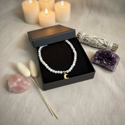 Maansteen ketting met kleine kralen en goudkleurige maan hanger in een sieradendoosje naast kristallen en kaarsen