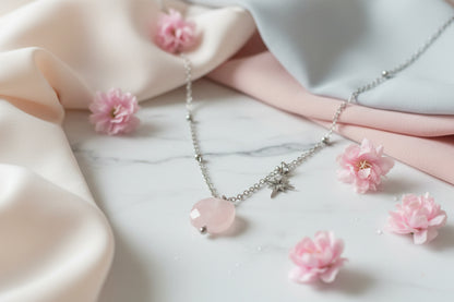 Luxe productfoto van de zilverkleurige RVS ketting met Rozenkwarts edelsteen en ster-charm op een marmeren ondergrond met roze bloemen eromheen - Glow Mystic in Style.