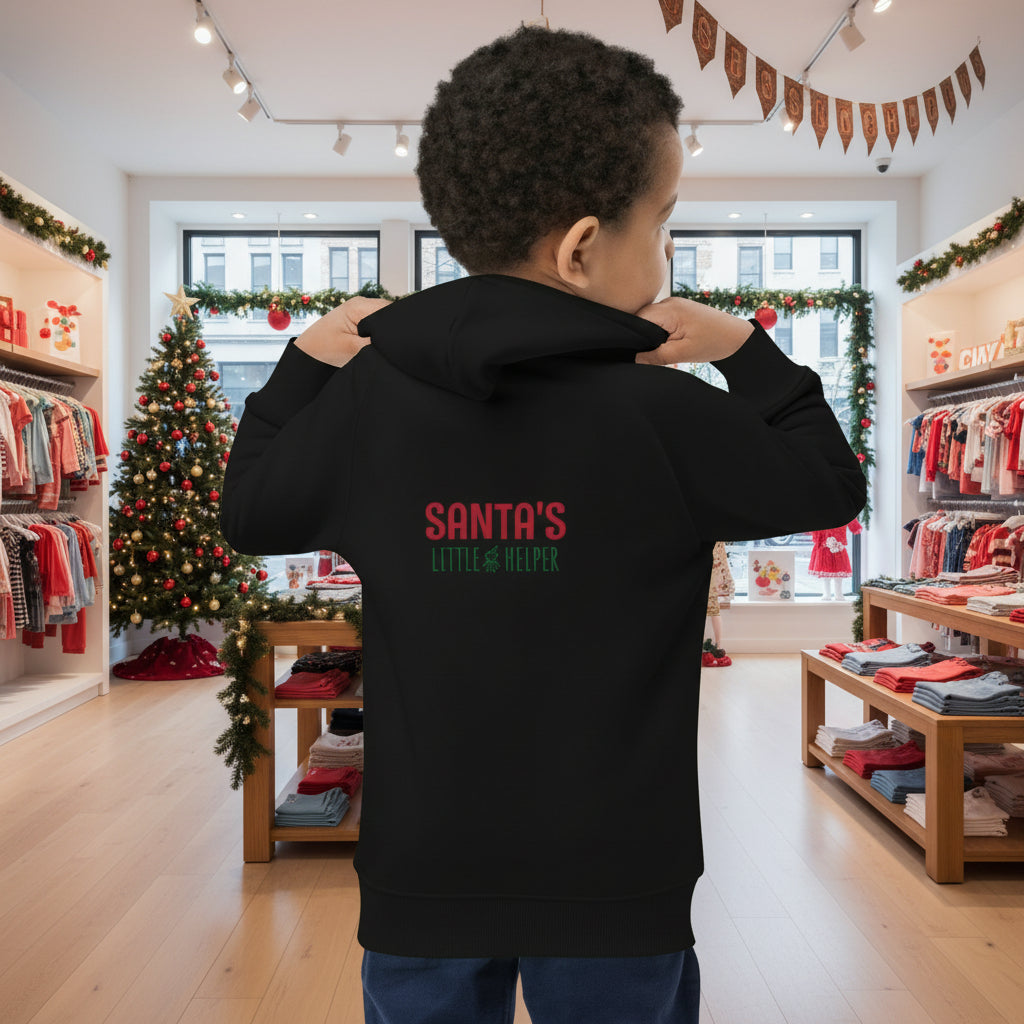 Child wearing black Santa's Little Helper Kids Hoodie with festive text by Christmas tree indoors