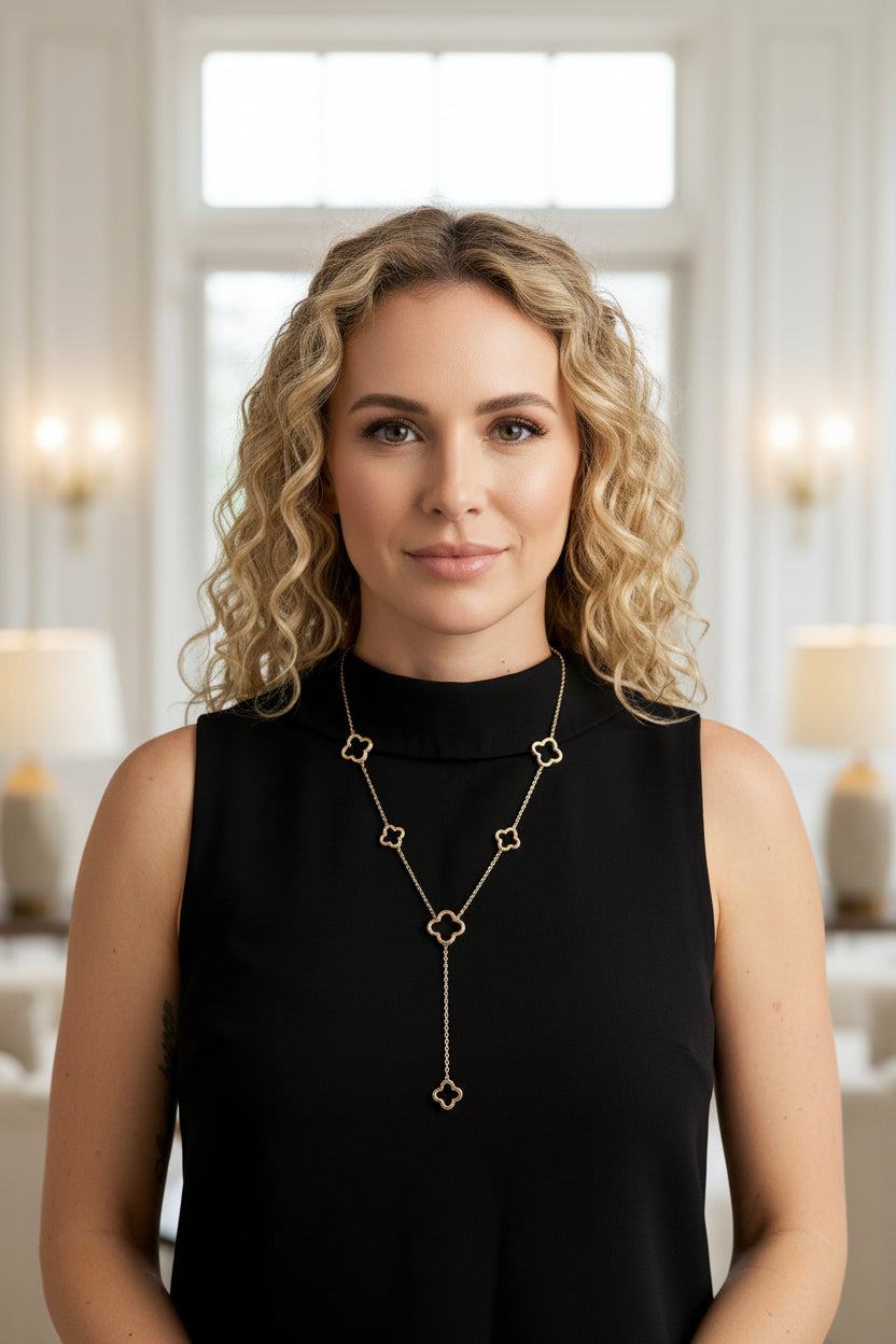 Gestileerd portret van een vrouw met krullend blond haar en een zwarte top, die de goudkleurige klaver lariat-ketting draagt, waardoor de verfijnde 'open-work' klavers en de elegante hanger prachtig over haar halslijn vallen."