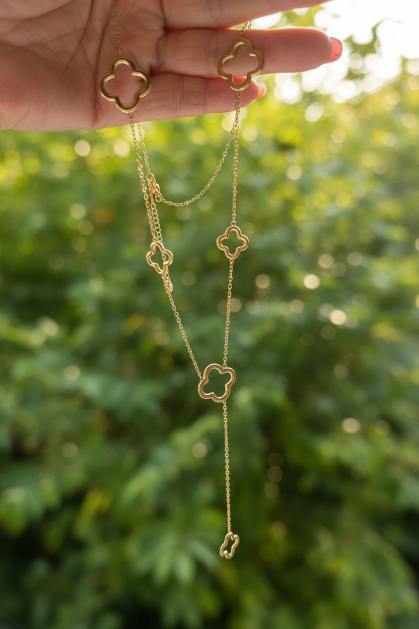 "Sfeerfoto van de goudkleurige klaver lariat-ketting, teder vastgehouden in een hand met roze glitter nagellak, in een zonnige, natuurlijke buitenomgeving met vervaagd groen."