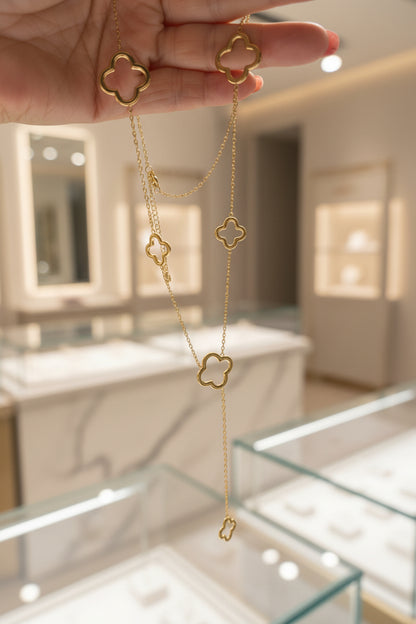 Een goudkleurige , gelaagde ketting met meerdere open klavertjevier elementen van verschillende groottes, omhooggehouden in een hand tegen de lichte, wazige achtergrond van een luxe sieradenboetiek met verlichte vitrines.