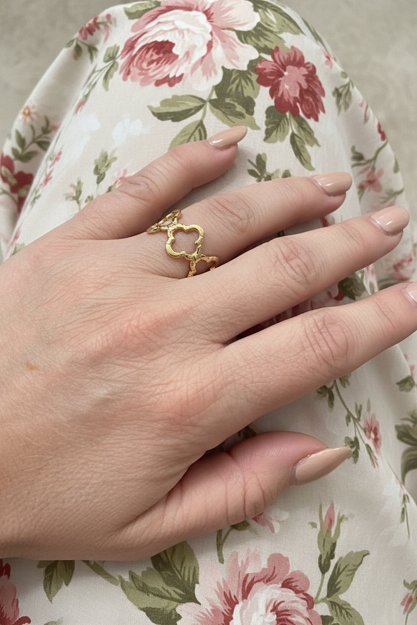 "Goudkleurige RVS klaver ring gedragen aan de hand tegen een achtergrond van een bloemenprint jurk, luxe en vrouwelijke uitstraling."