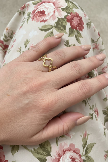 "Goudkleurige RVS klaver ring gedragen aan de hand tegen een achtergrond van een bloemenprint jurk, luxe en vrouwelijke uitstraling."