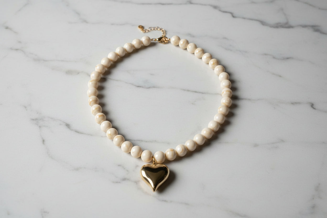 Close-up van de gepolijste goudkleurige harthanger en de beige-witte gevlamde kralen van de Stainless Steel ketting op een marmeren achtergrond.