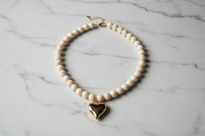 Close-up van de gepolijste goudkleurige harthanger en de beige-witte gevlamde kralen van de Stainless Steel ketting op een marmeren achtergrond.