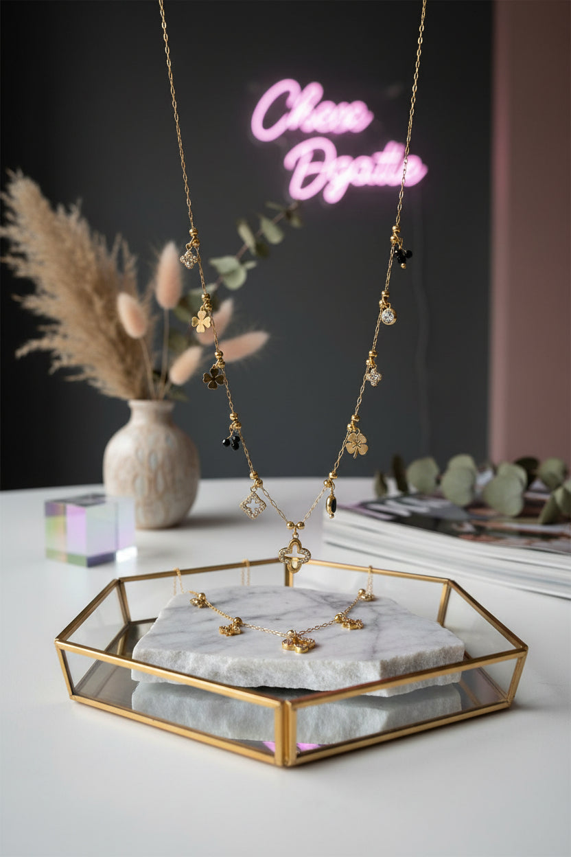 Een goudkleurige, gelaagde ketting met diverse fijne ruitvormige en geometrische hangers, elegant gedrapeerd op een wit marmeren dienblad met op de achtergrond zachtroze rozenblaadjes.