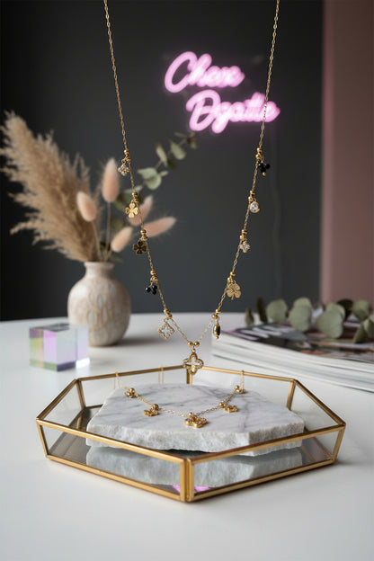 Een goudkleurige, gelaagde ketting met diverse fijne ruitvormige en geometrische hangers, elegant gedrapeerd op een wit marmeren dienblad met op de achtergrond zachtroze rozenblaadjes.