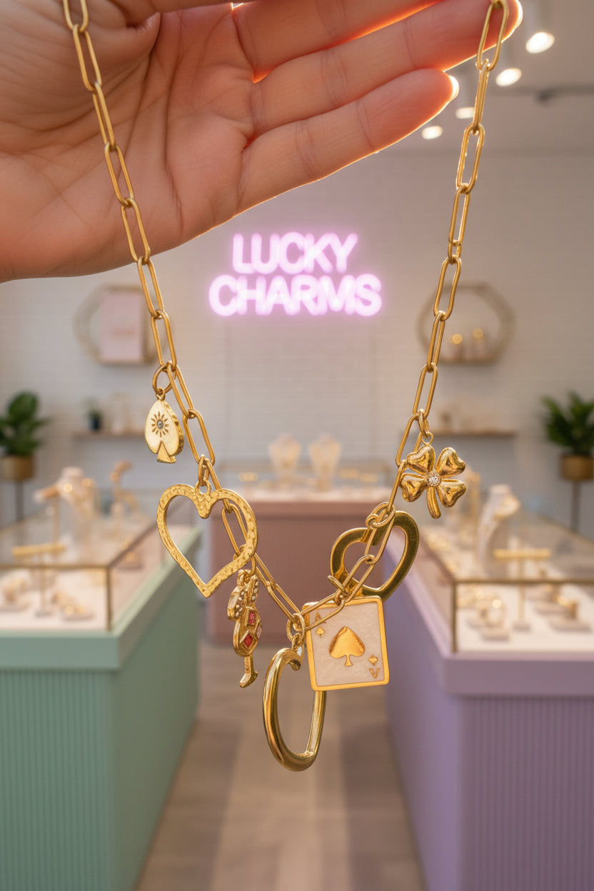 Close-up van een hand die een goudkleurige grove schakelketting omhoog houdt met diverse bedels, waaronder een groot hart, een klavertje vier, een speelkaart en een cocktailglas, tegen de wazige achtergrond van een luxe sieradenwinkel met een roze neonreclame waarop Lucky Charms staat.