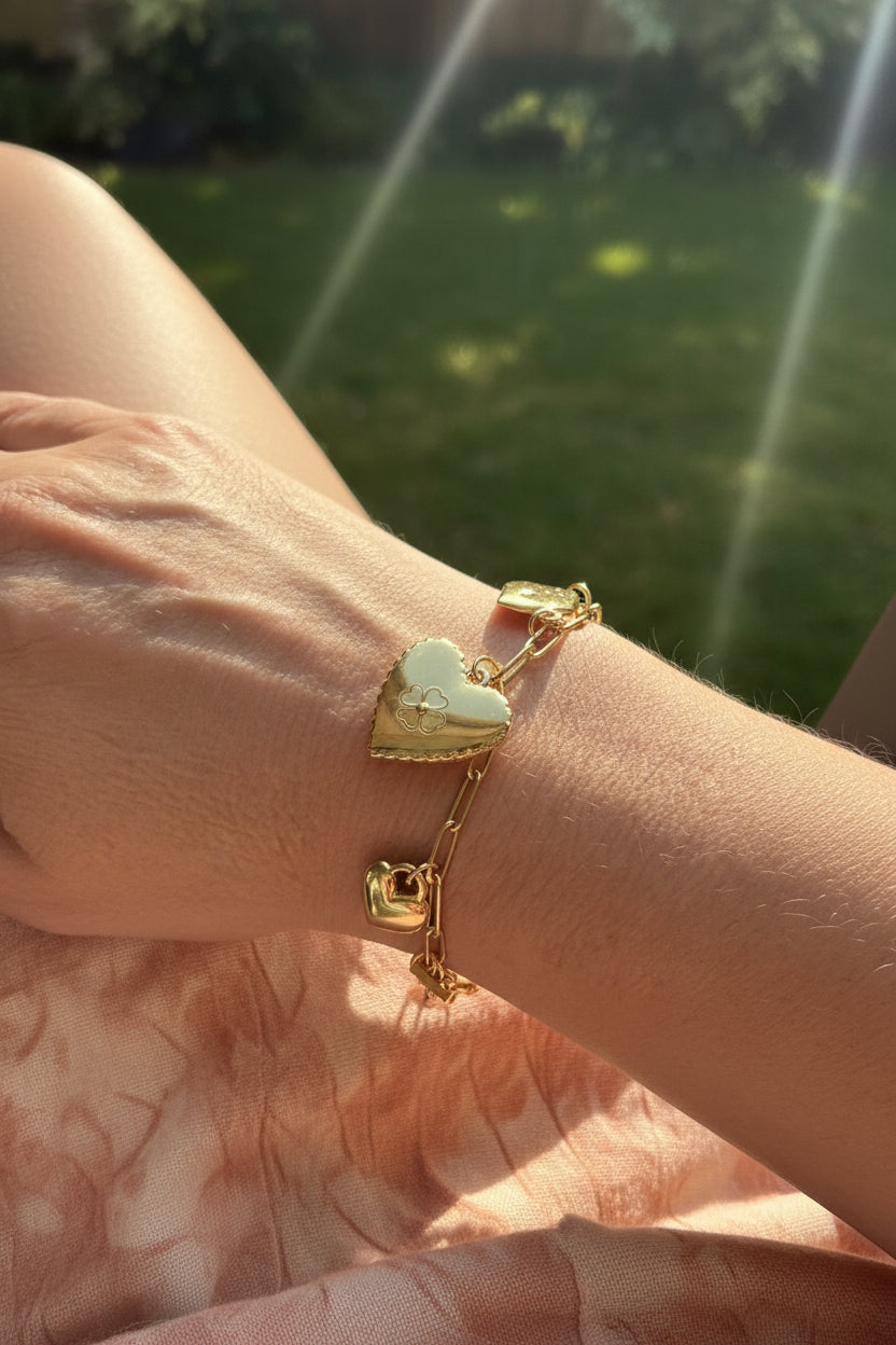 Goudkleurige RVS bedelarmband met een groot hartje en klavertje vier, elegant gedragen om de pols van een model in de tuin.
