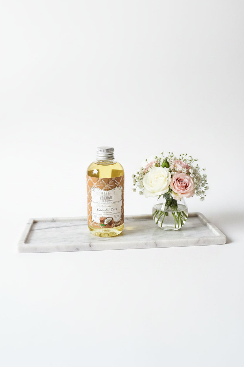 Saules Fabrika massageolie Coconut 200ml op een luxe marmeren plateau naast een vaasje met zachtroze en witte rozen - Glow Mystic in Style.