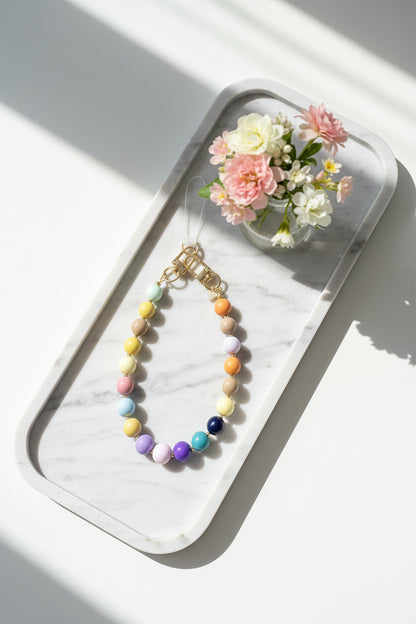 Multicolor telefoonkoord met pastelkleurige kralen en goudkleurige sluiting, stijlvol gepresenteerd op een marmeren dienblad naast een klein bloemstuk.