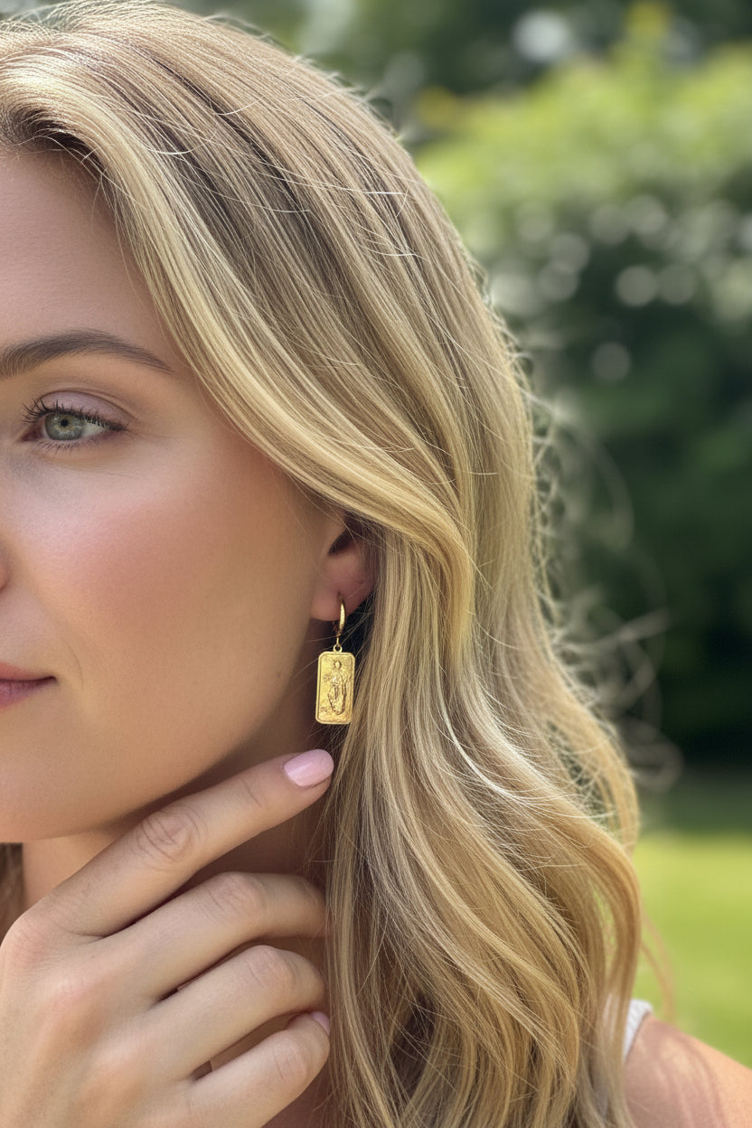 Een blond model met zachtroze nagellak draagt de goudkleurige RVS mystic hand statement oorbel in een zonnige buitenomgeving.