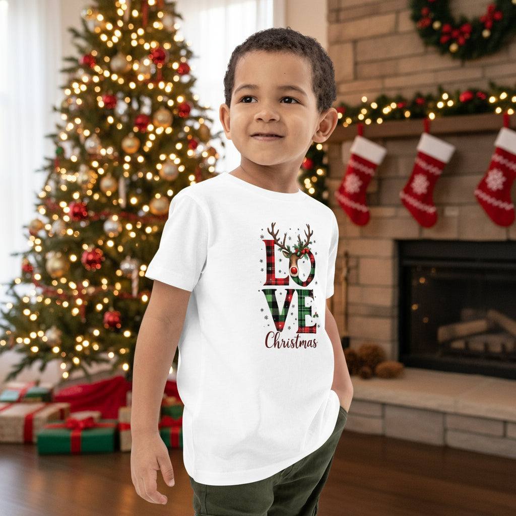 Child wearing white organic kids T-shirt with festive Love Christmas design by decorated Christmas tree and fireplace