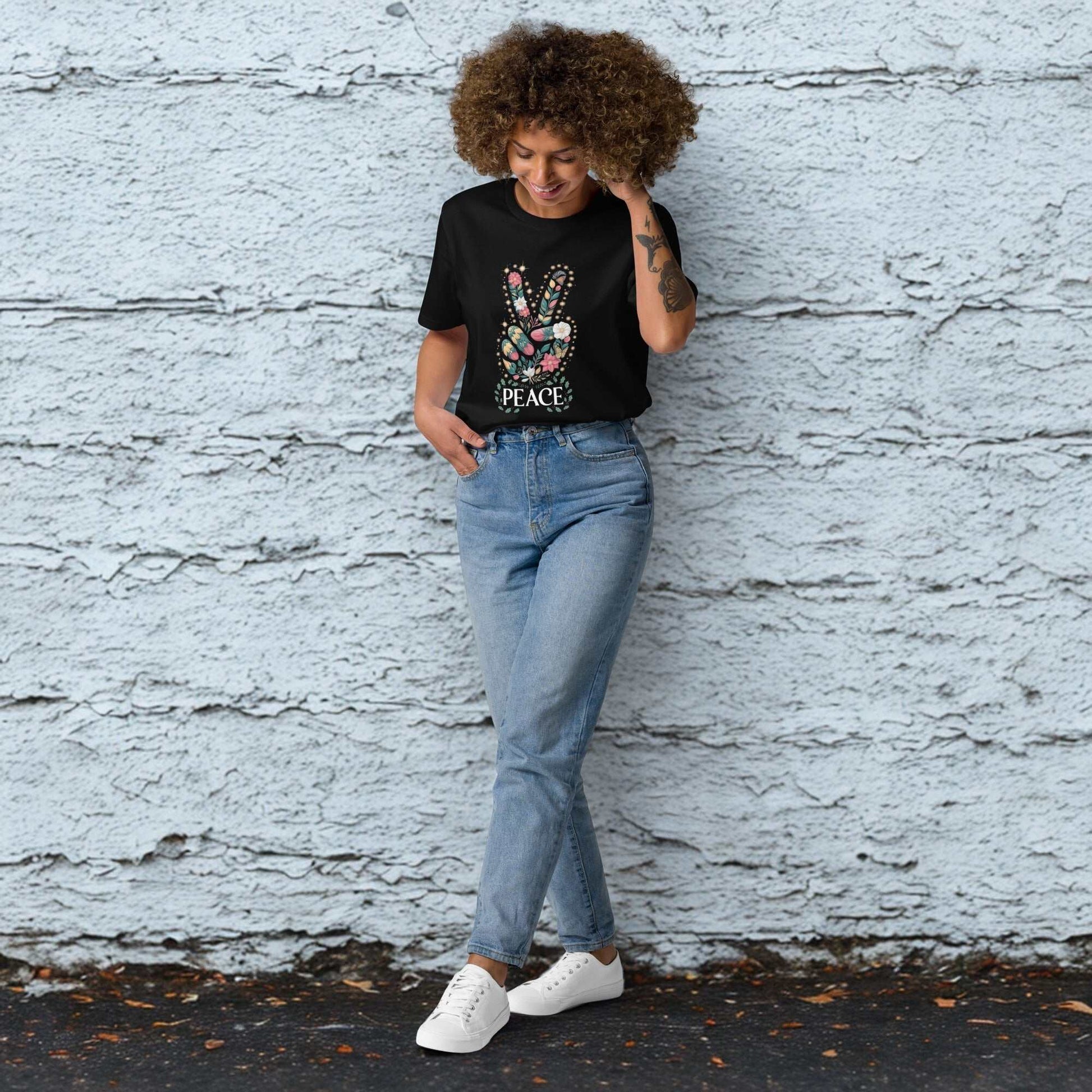 Woman wearing a black unisex organic cotton T-shirt with a peace sign graphic, paired with blue jeans and white sneakers, standing against a textured gray wall