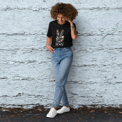 Woman wearing a black unisex organic cotton T-shirt with a peace sign graphic, paired with blue jeans and white sneakers, standing against a textured gray wall