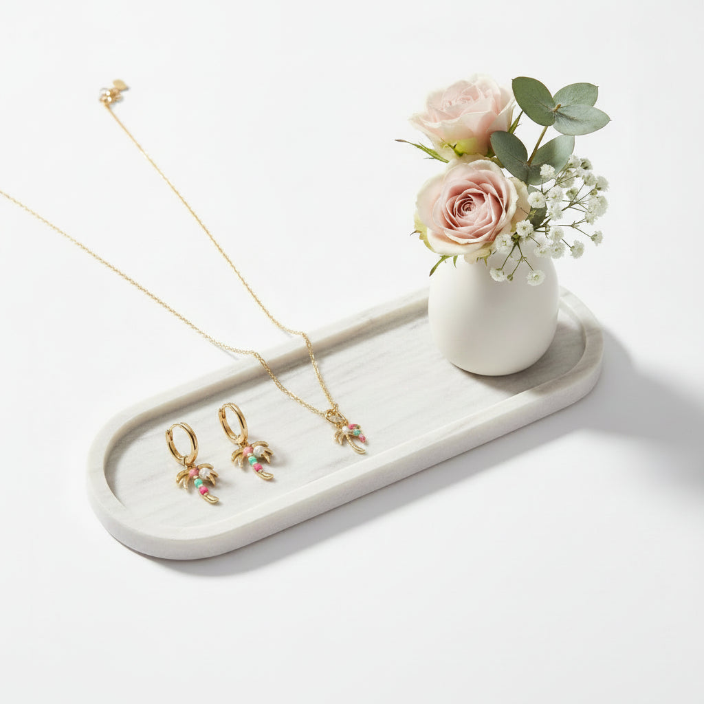 Een goudkleurige sieradenset bestaande uit een fijne ketting en bijpassende creolen oorbellen met palmhangers, versierd met gekleurde steentjes, gepresenteerd op een wit marmeren tray naast een klein wit vaasje met zachtroze rozen en gipskruid.