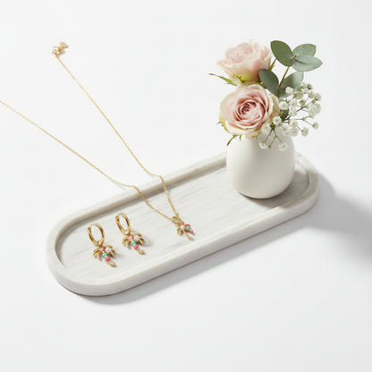Een goudkleurige sieradenset bestaande uit een fijne ketting en bijpassende creolen oorbellen met palmhangers, versierd met gekleurde steentjes, gepresenteerd op een wit marmeren tray naast een klein wit vaasje met zachtroze rozen en gipskruid.