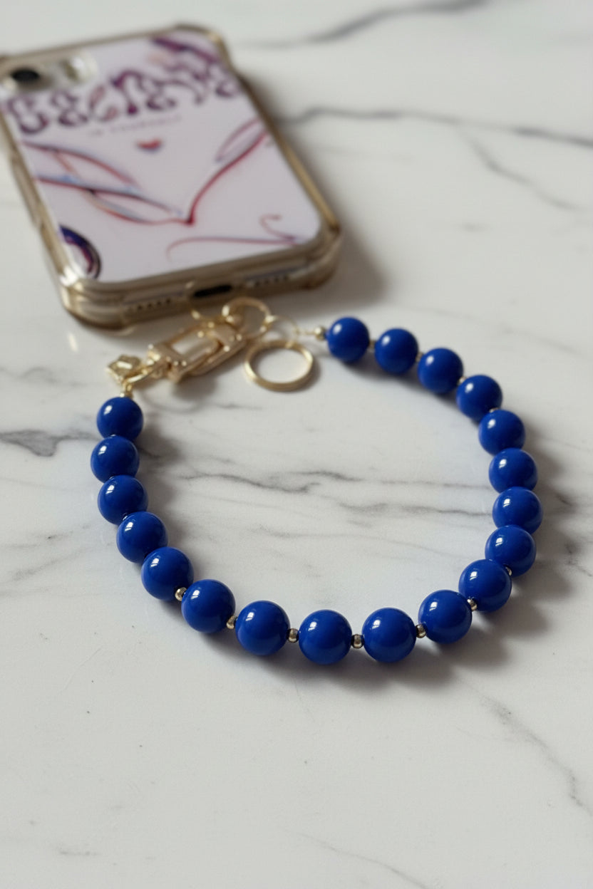 Handgemaakte phone charm met koningsblauwe kralen en gouden details, gefotografeerd op een luxe witte marmeren achtergrond.