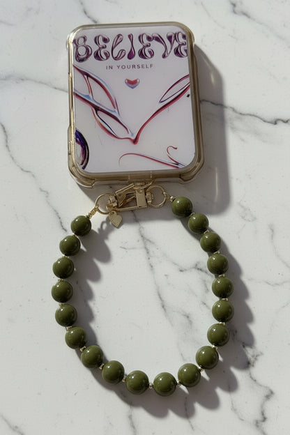 "Lifestyle foto van de Olive Green phone charm bevestigd aan een hoesje met de tekst 'Believe in yourself'."