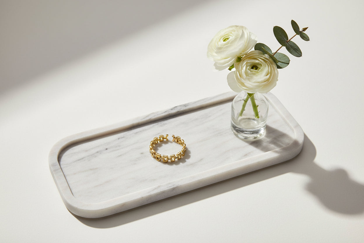 Een verstelbare gouden ring met een open schakel - of kettingschakelontwerp, liggend op een langwerpig wit dienblad naast een klein glazen vaasje met twee witte ranoenkels en een takje eucalyptus.