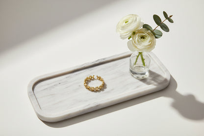 Een verstelbare gouden ring met een open schakel - of kettingschakelontwerp, liggend op een langwerpig wit dienblad naast een klein glazen vaasje met twee witte ranoenkels en een takje eucalyptus.