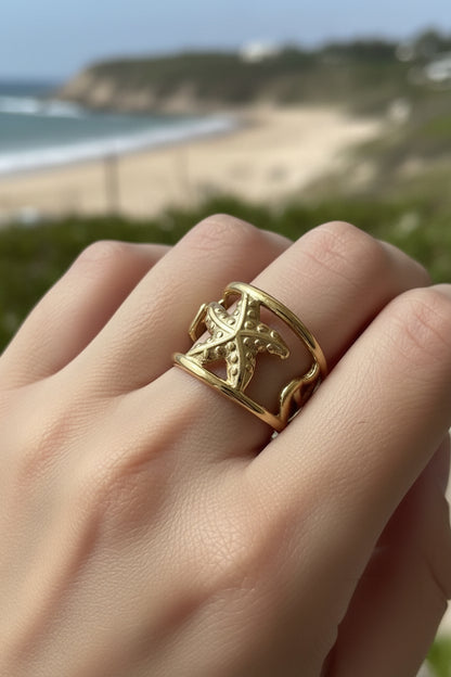 Detailopname van een hand die een brede goudkleurige zeester ring draagt, met een zomers strand en de zee op de achtergrond.