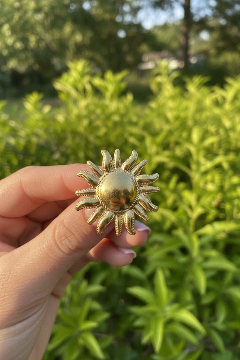 "Goudkleurige XL statement ring in de vorm van een zon, vastgehouden tegen een groene natuurlijke achtergrond."