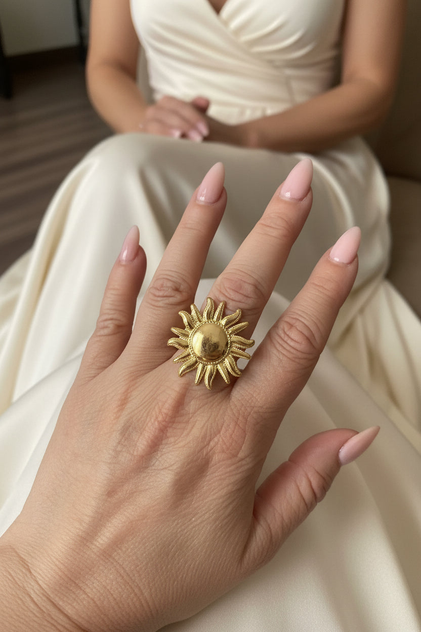 "Close-up van de XL gouden zon ring gedragen aan de hand van een vrouw in een elegante witte jurk."