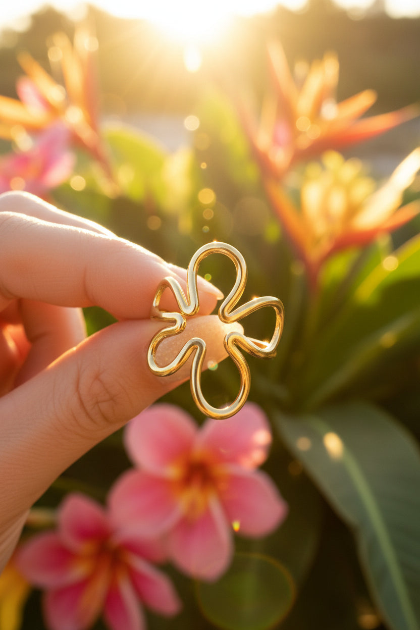"Sfeerfoto van de goudkleurige RVS Abstract Flower ring van Glow Mystic in Style, vastgehouden tegen een achtergrond van tropische bloemen in het zonlicht."