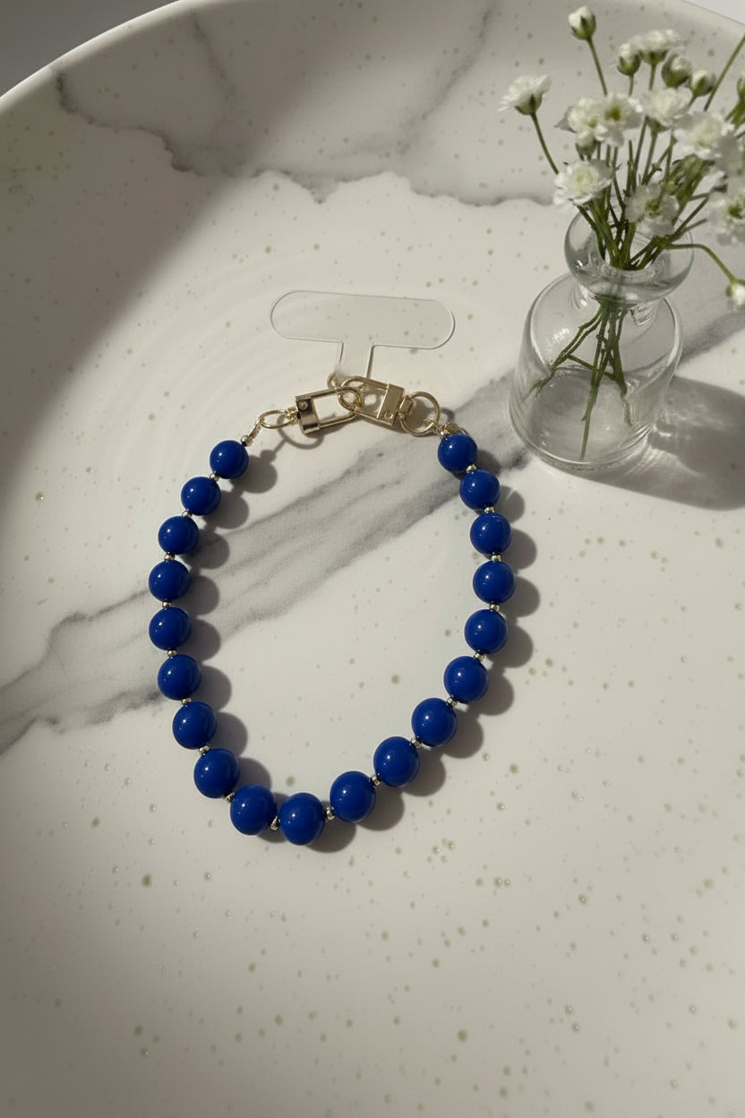 Handgemaakte phone charm met grote koningsblauwe acryl kralen en goudkleurige details op een marmeren schaal met een vaasje met bloemetjes ernaast .